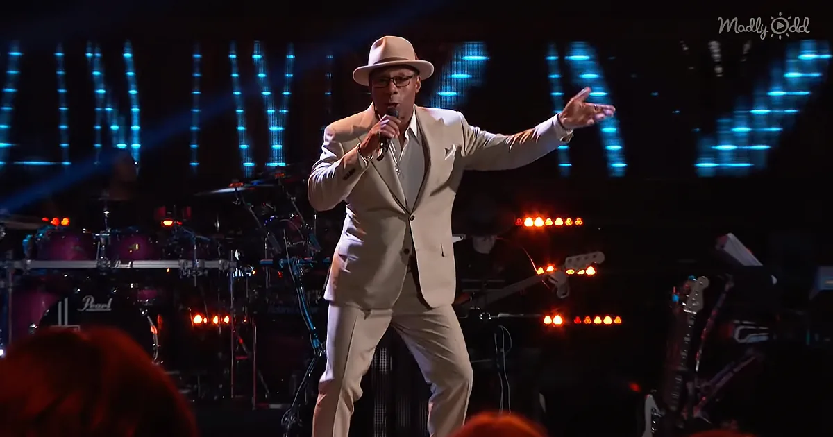 A man in a white suit and hat sings into a microphone on stage, with a band and blue lights in the background.