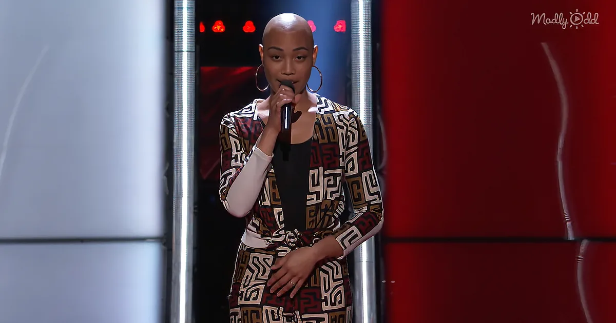 A person with a shaved head and large hoop earrings holds a microphone on stage, wearing a patterned outfit.