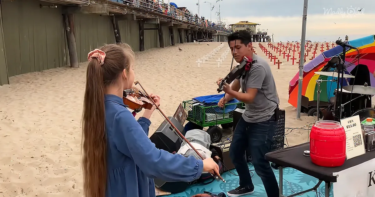 A young girl and boy play violins on a beach, with a colorful umbrella and drum set nearby.