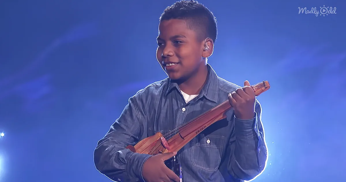 A young boy in a denim shirt plays a stringed instrument on stage under blue lighting.