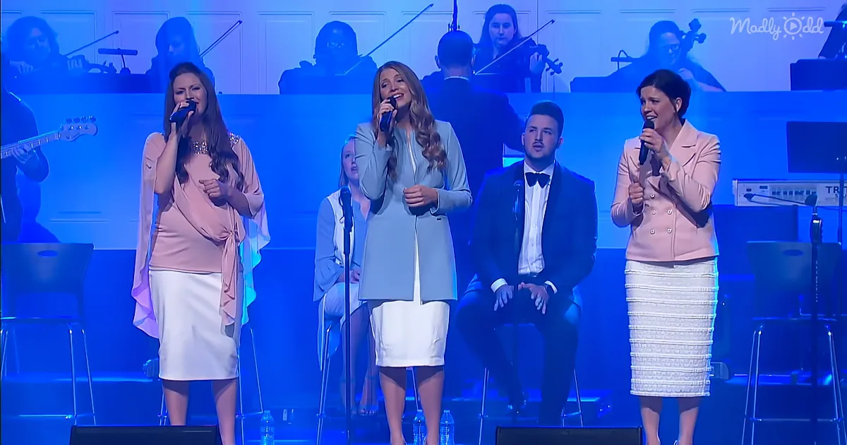 A group of four performers sing on stage with a musician and orchestra in the background.