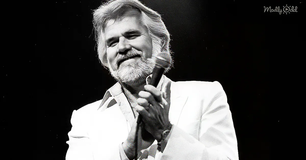 A man with a beard and mustache, wearing a white suit, holds a microphone and smiles on stage.