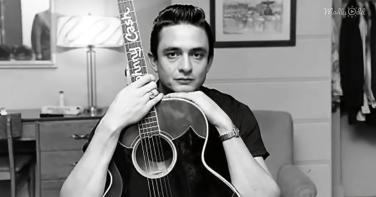 A man holds a guitar, wearing a black shirt, in a room with a lamp and framed pictures.