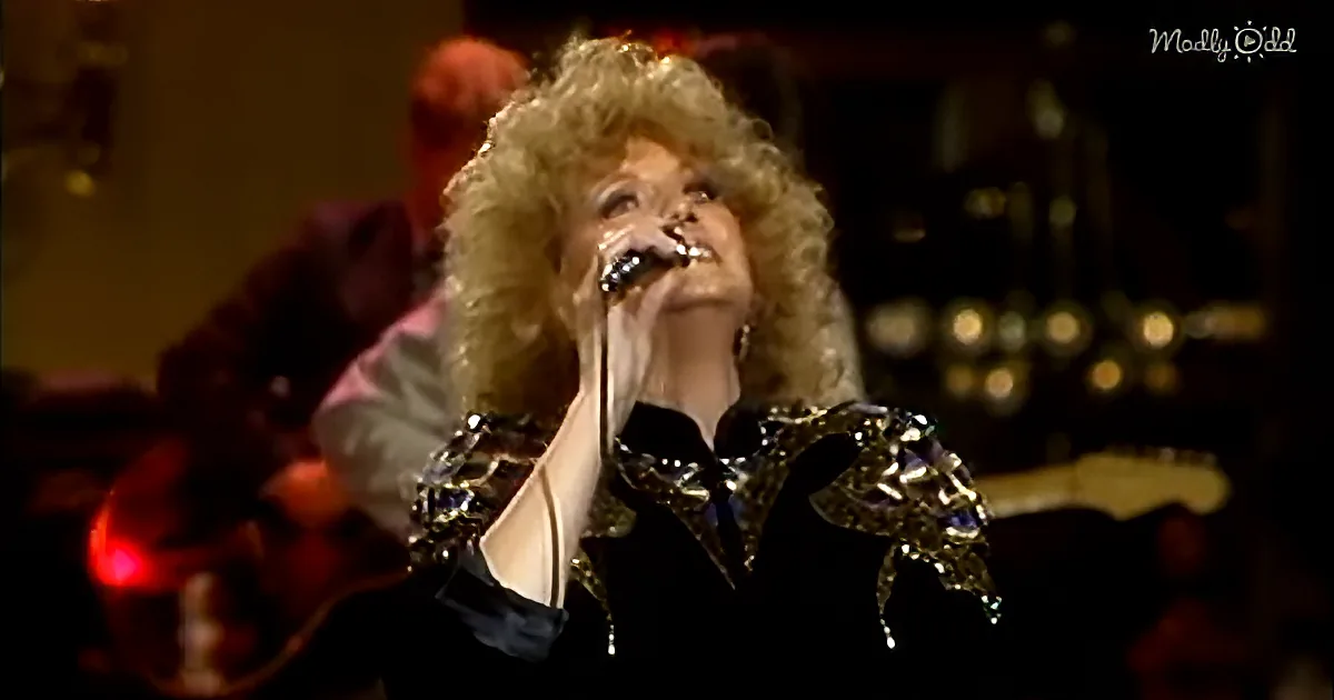 A woman with curly blonde hair sings into a microphone, wearing a black outfit with gold embellishments.