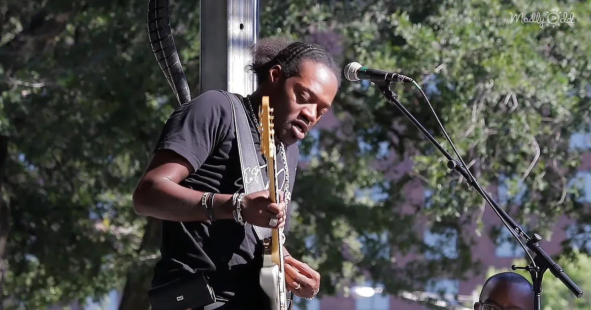 A man plays guitar under a microphone, surrounded by trees and sunlight.