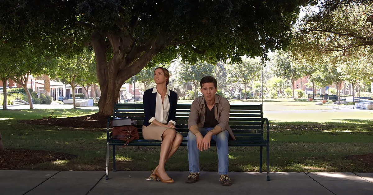A man and woman sit on a bench in a park, surrounded by trees and greenery.