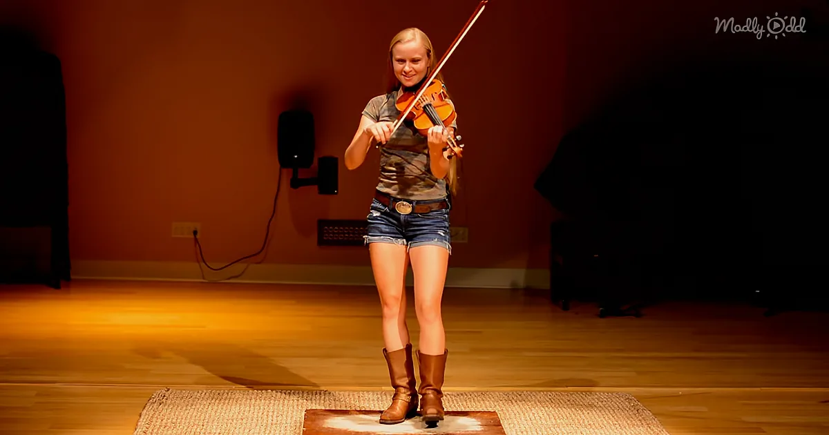A person plays violin on stage, wearing denim shorts and boots, with a spotlight on them.