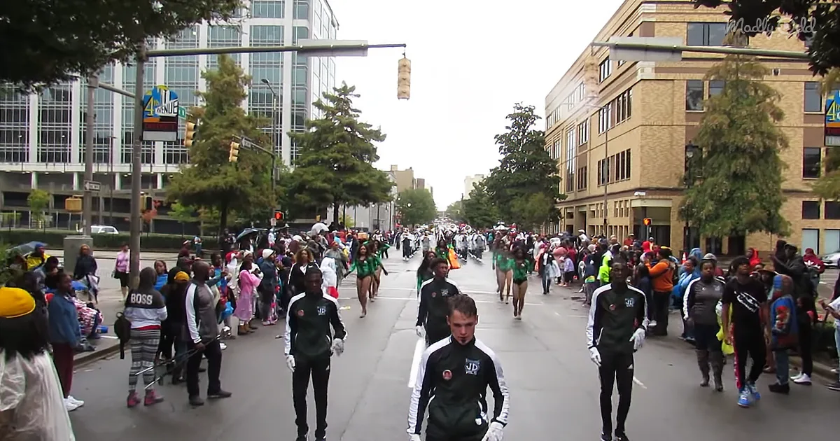 Marching band in black uniforms leads a parade down a city street with spectators on both sides.