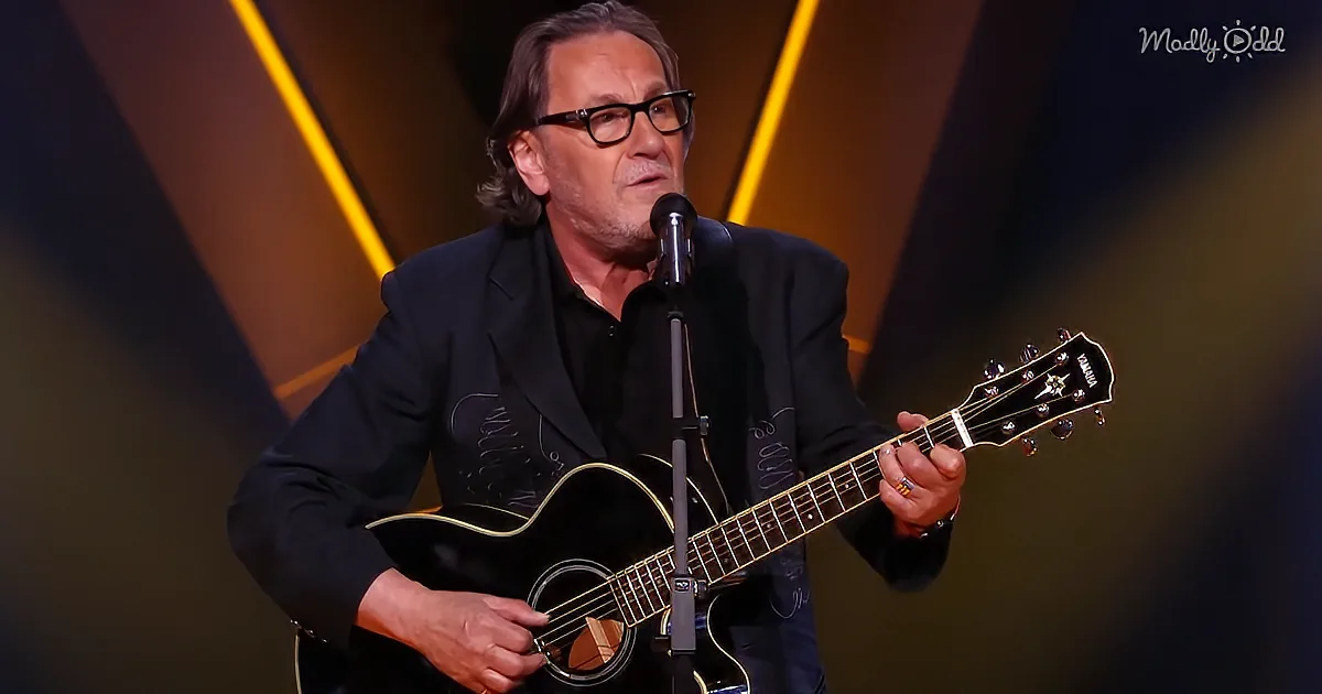A man in a black suit plays a black acoustic guitar on stage with a microphone.