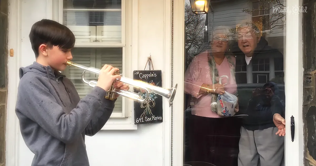A boy plays trumpet outside a house, while an elderly couple looks on from inside.