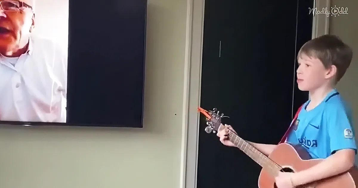 A young boy plays guitar while a man sings on a TV screen behind him.