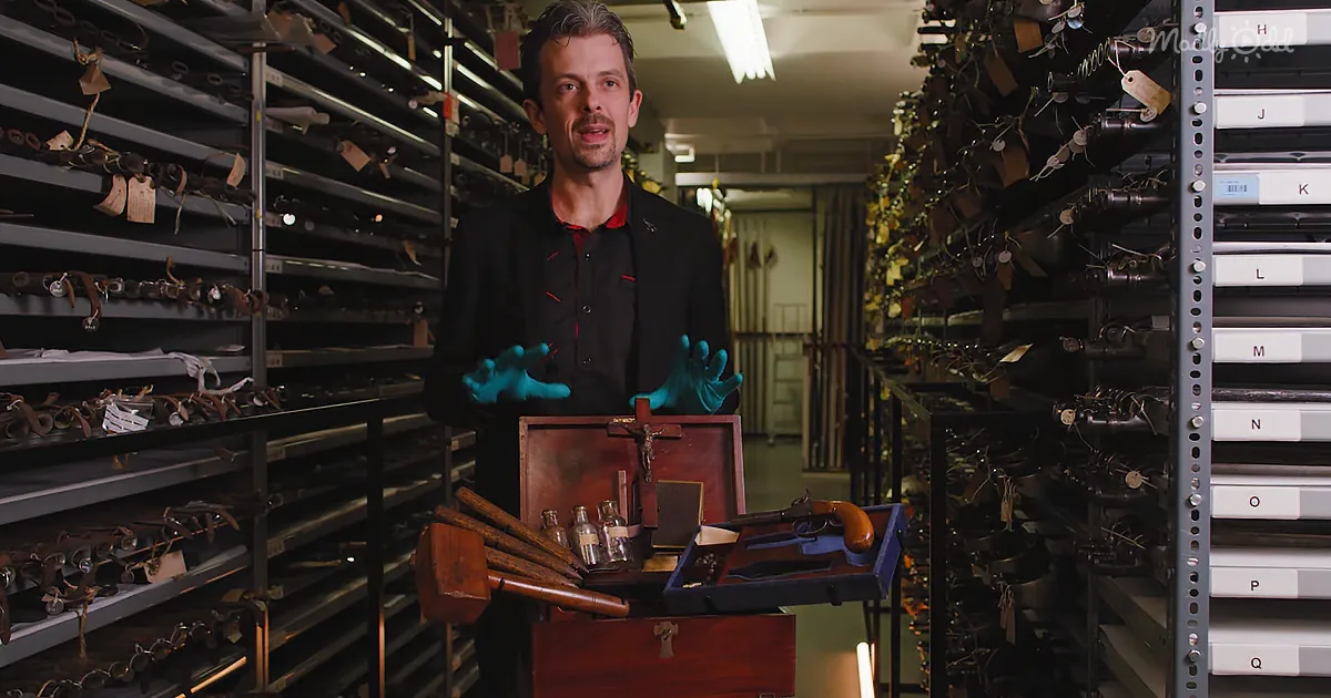 A man in a black suit and blue gloves stands in a room filled with shelves of weapons and tools.