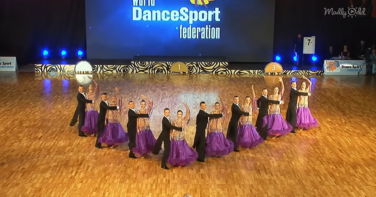 A group of dancers in purple dresses and black suits pose on a wooden floor under bright stage lights.