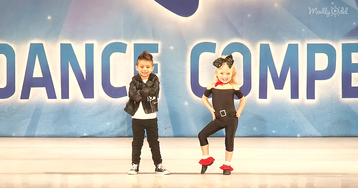 A boy and girl stand on stage in front of a "DANCE COMPETITION" backdrop, both dressed in dance attire.