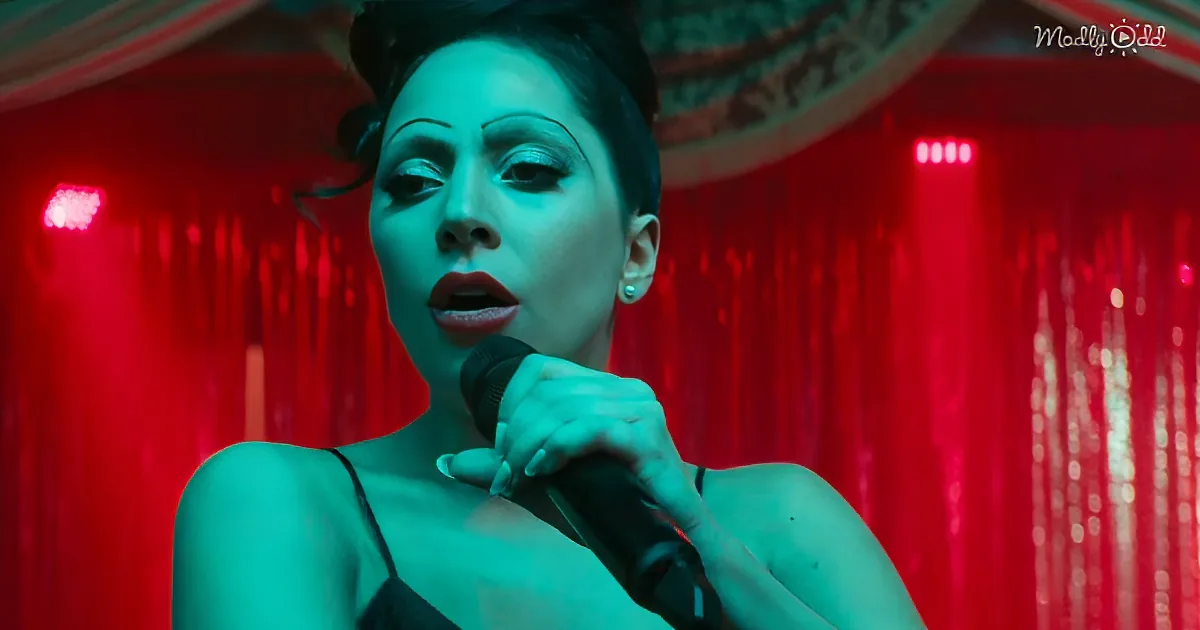 A woman with dark hair and makeup holds a microphone, singing in front of red curtains.