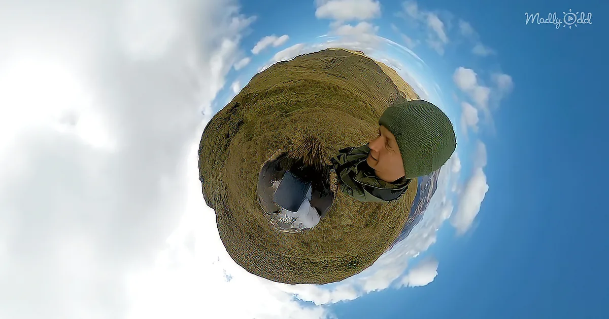 A person wearing a green beanie and jacket stands on a grassy hill under a blue sky with clouds.