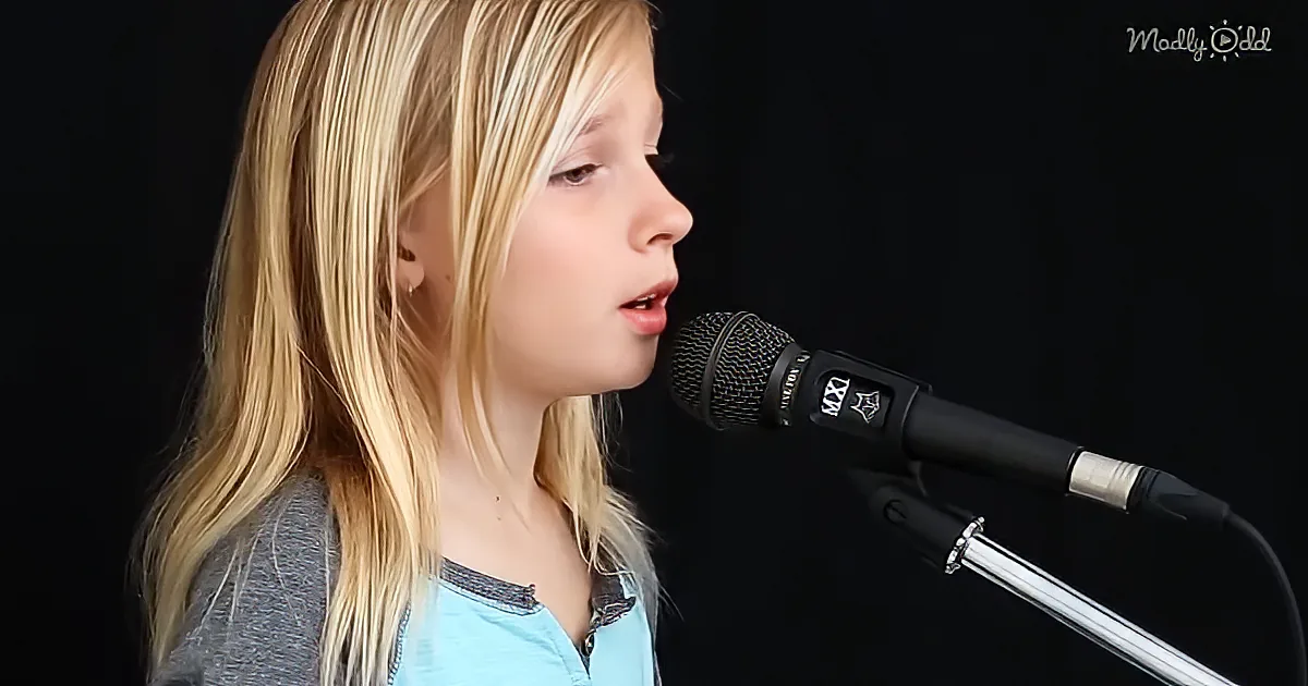 A young girl with long blonde hair sings into a microphone against a black background.