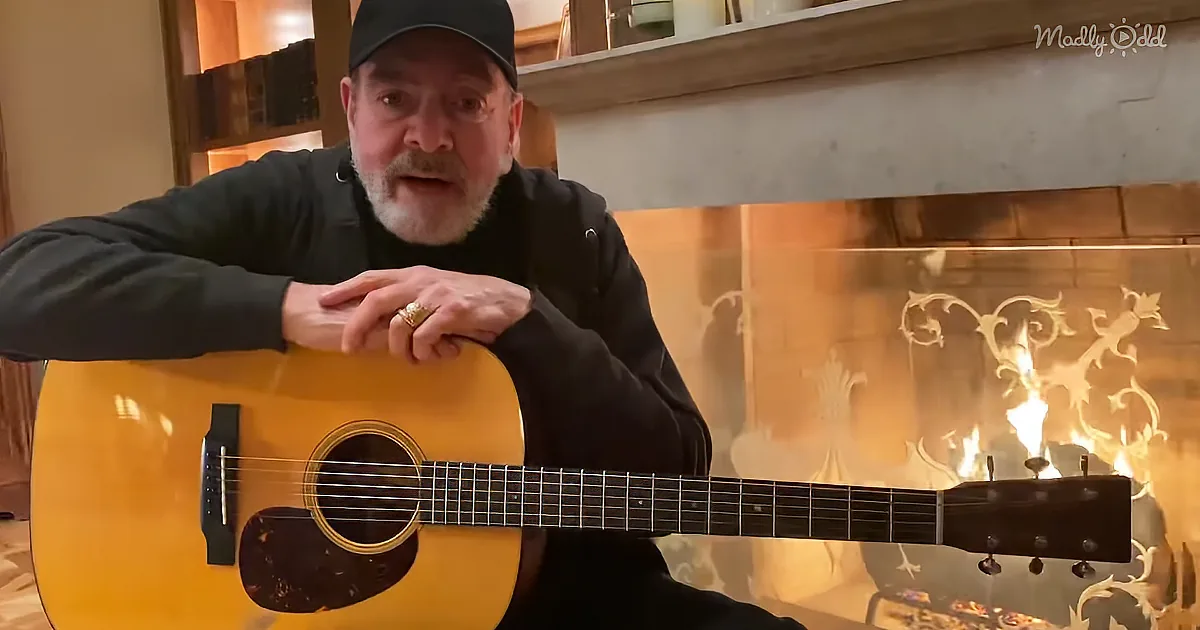 Man in black shirt and cap plays acoustic guitar in front of fireplace with flames.