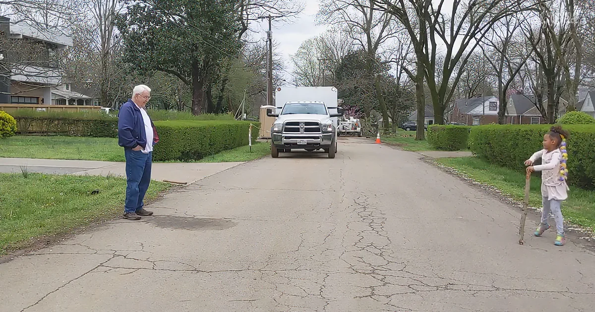 An elderly man stands on a cracked driveway, watching a white truck drive by in a suburban neighborhood.