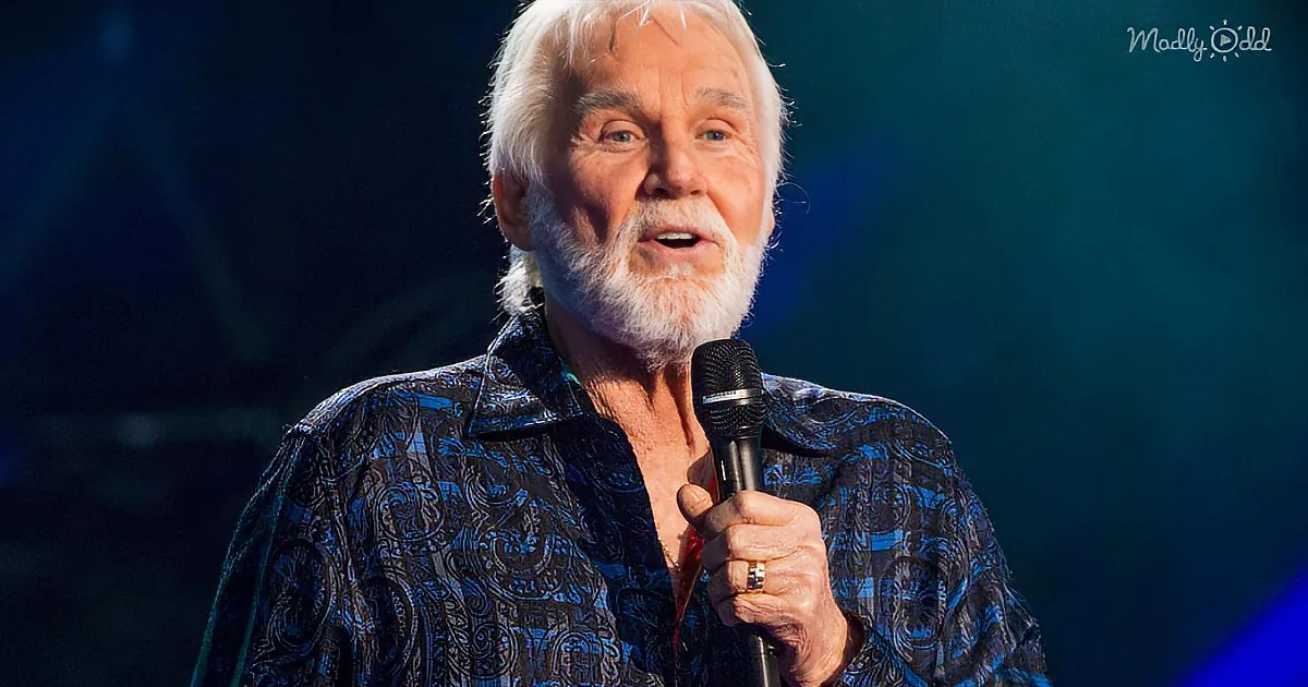 Kenny Rogers holds a microphone, wearing a patterned shirt, and appears to be singing or speaking on stage.