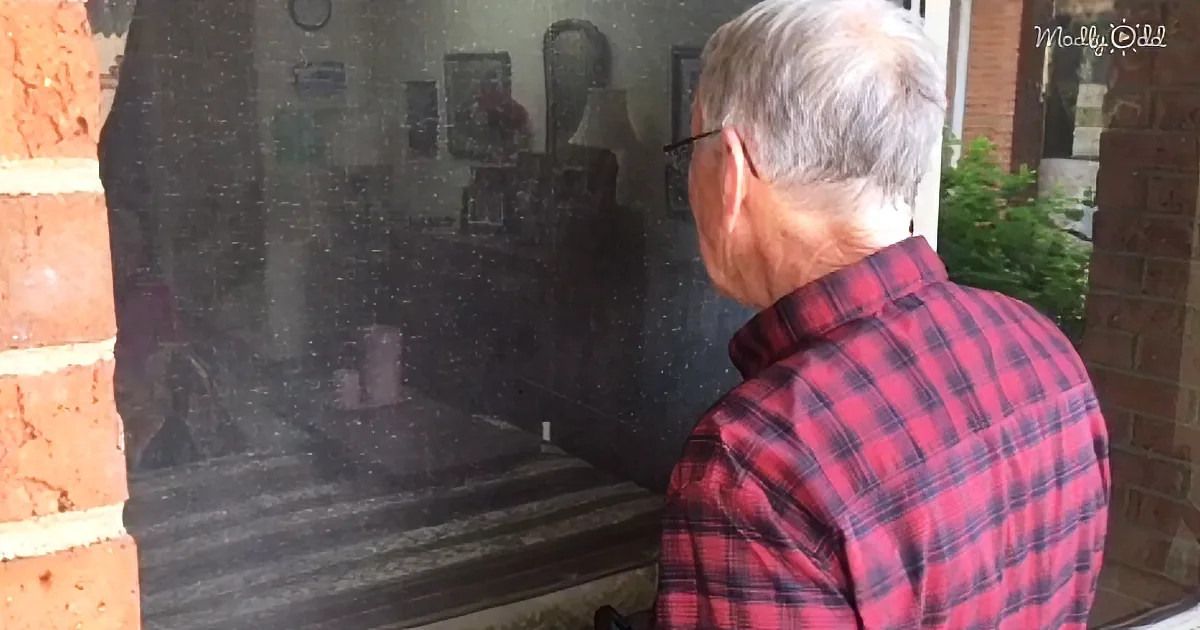 An older man in a red plaid shirt stands outside, looking into a glass door with reflections of furniture inside.