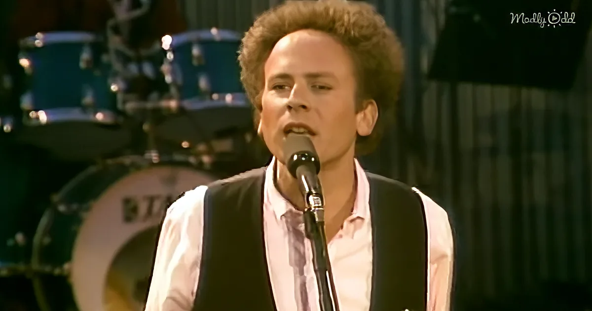 A person in a pink shirt and black vest sings into a microphone with a drum set in the background.