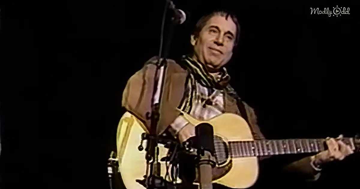 A man plays an acoustic guitar on stage, wearing a scarf, with a microphone in front of him.