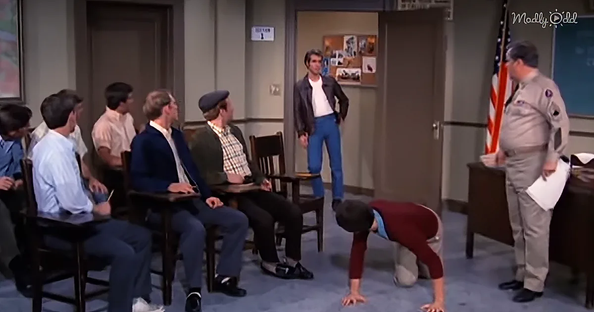 A man in a red shirt and khaki pants is doing a handstand in a room with seated men and a standing man in a uniform.