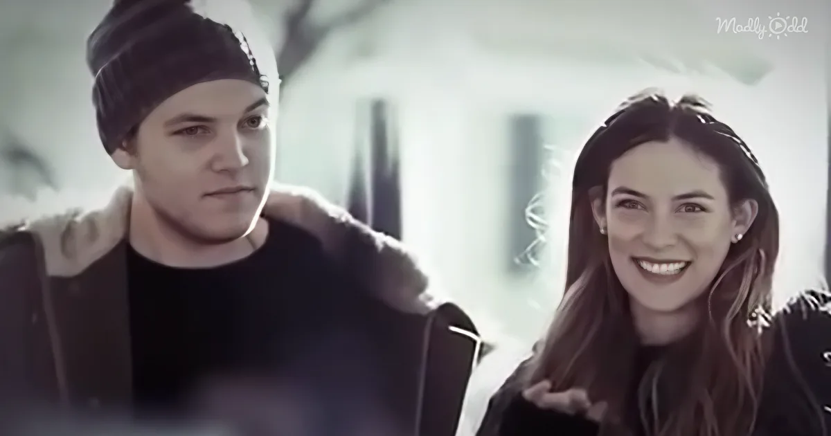 A man and woman are sitting together, the woman smiling and the man looking down, both in a casual setting.