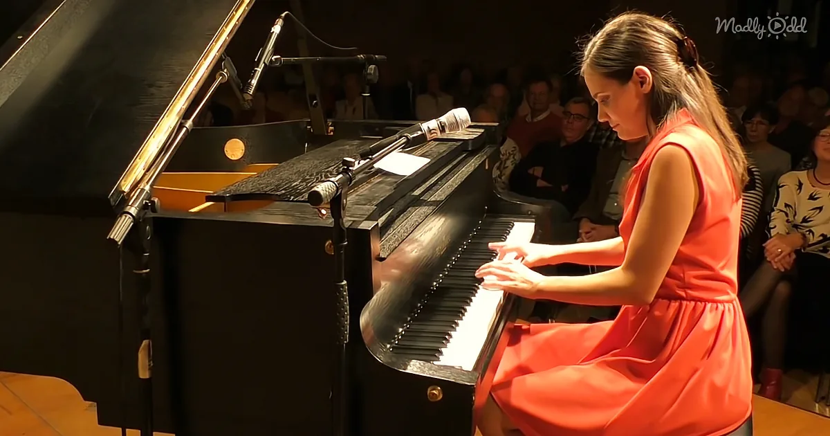 A woman in a red dress plays a grand piano in front of an audience.