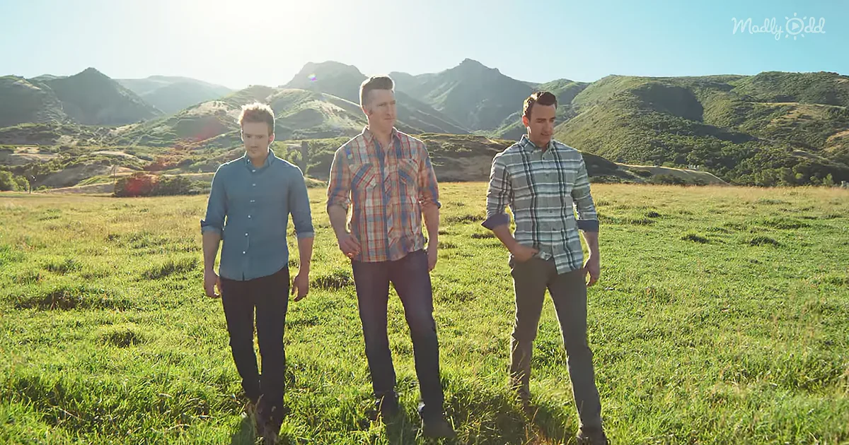Three men stand in a grassy field with mountains in the background under a bright sky.