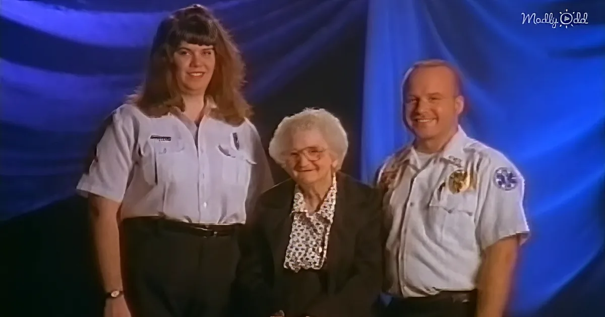 An elderly woman sits between two uniformed individuals on a stage with blue lighting.