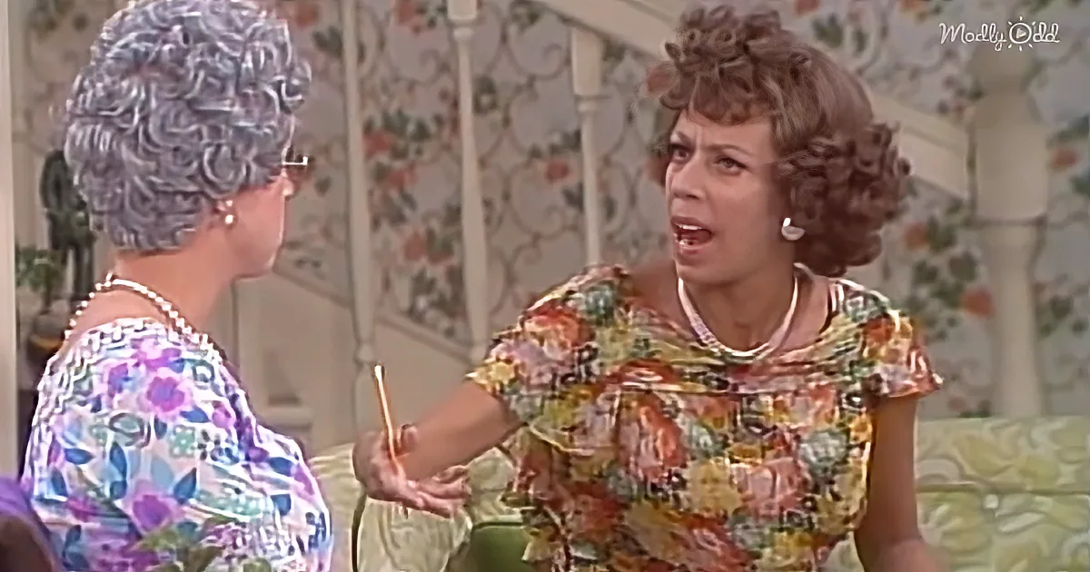 Woman in floral dress holding pencil, talking to another woman in floral top, both in a room with floral wallpaper.
