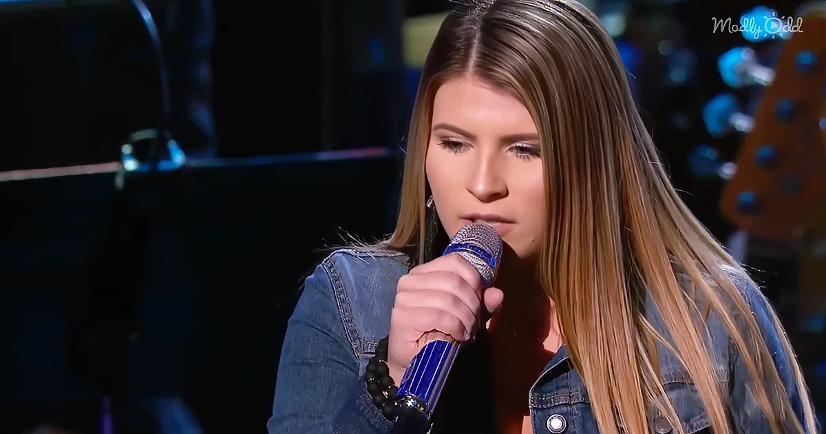 A woman with long hair holds a microphone, wearing a denim jacket, in a dimly lit setting.