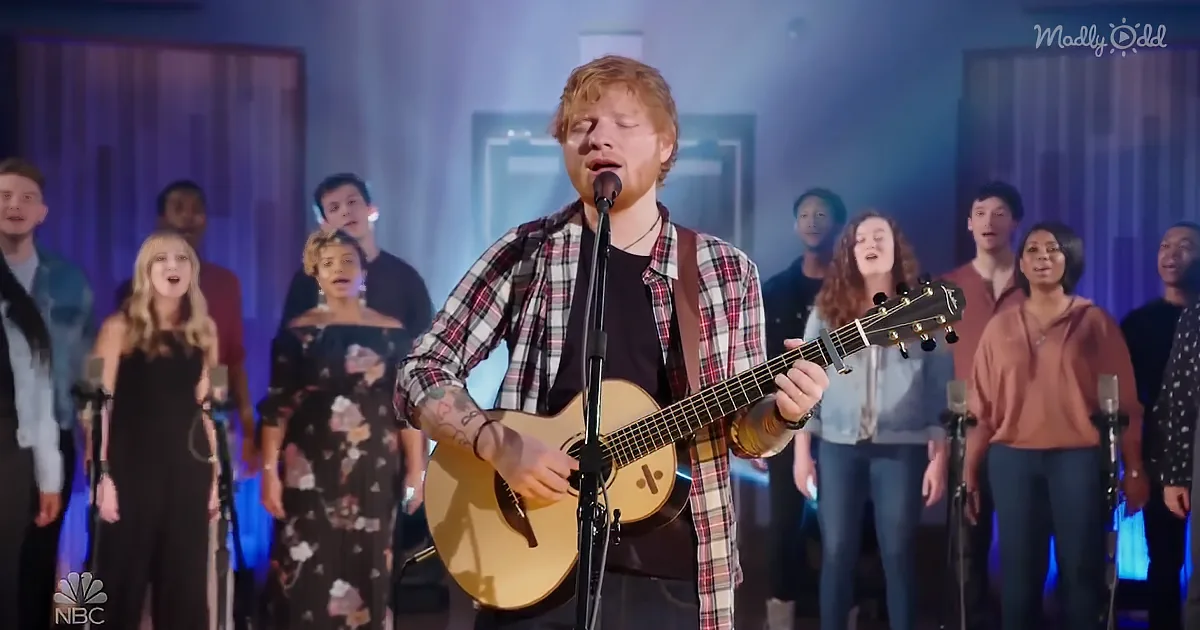 A man with red hair plays guitar and sings in front of a choir.