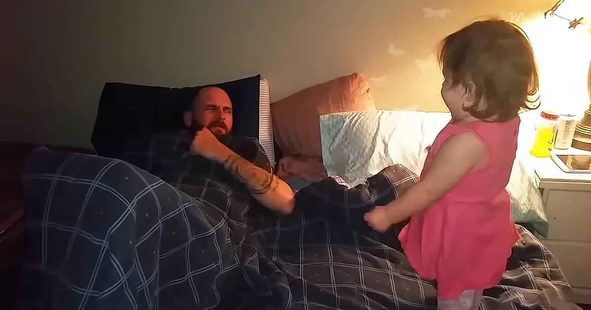 A man in bed with a child in a pink dress standing beside him, holding his leg.