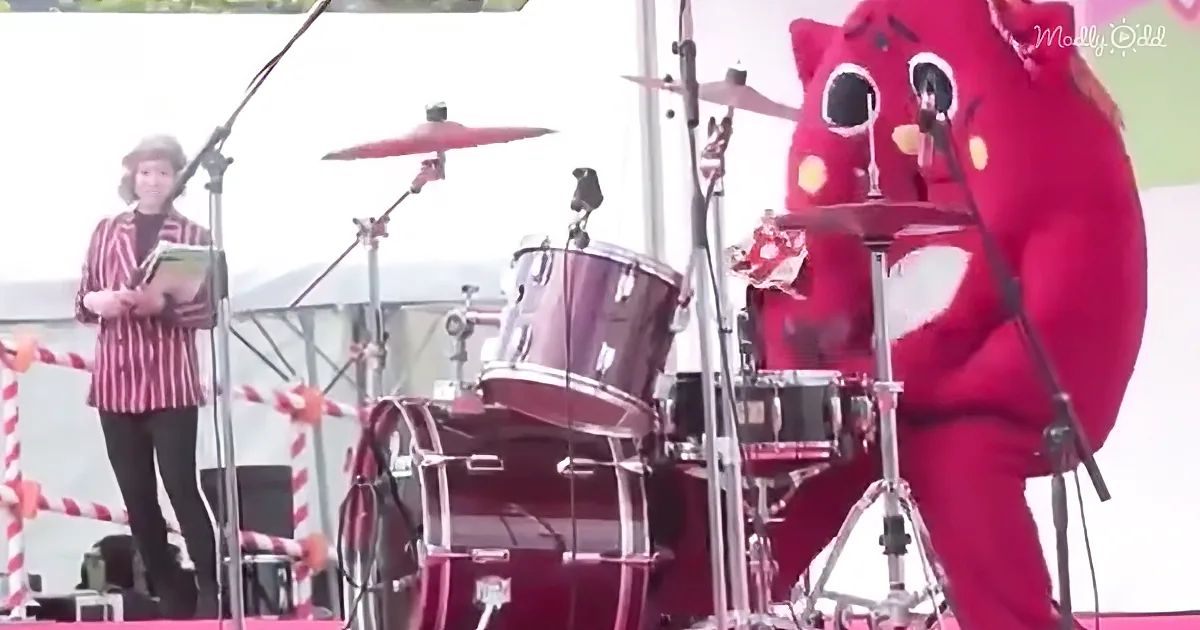 A person in a red costume plays drums on stage, while another person stands nearby holding a book.