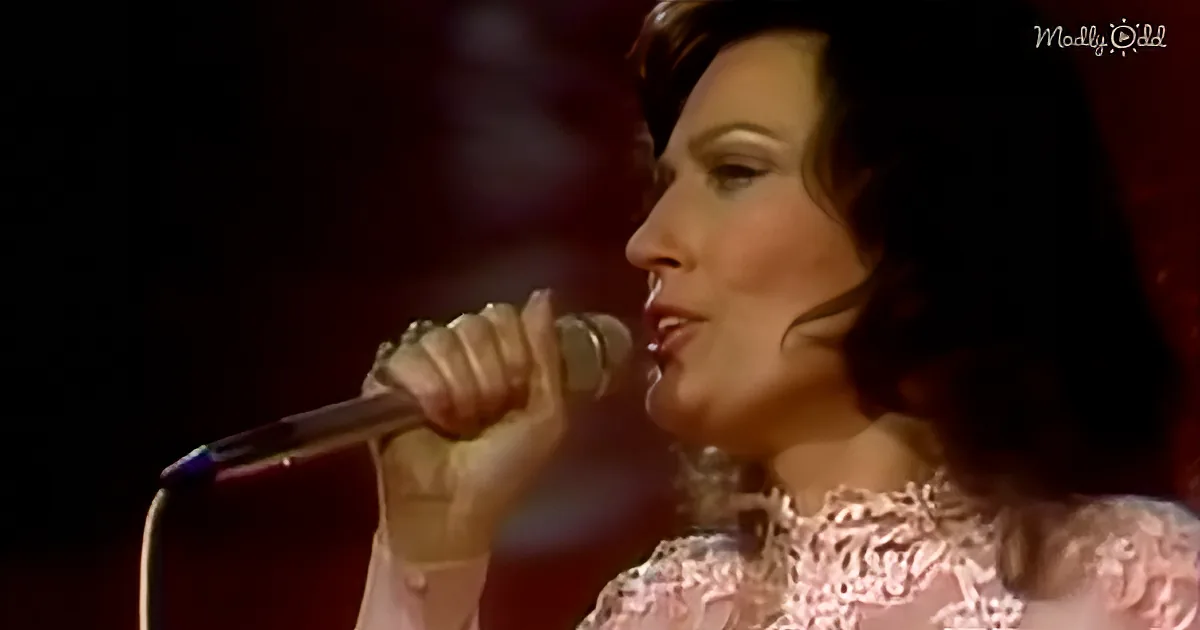 A woman in a pink dress sings into a microphone, holding it with her right hand.