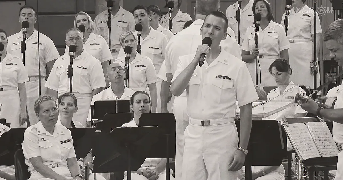 A man in white uniform sings into a microphone, surrounded by a choir and musicians in a formal setting.