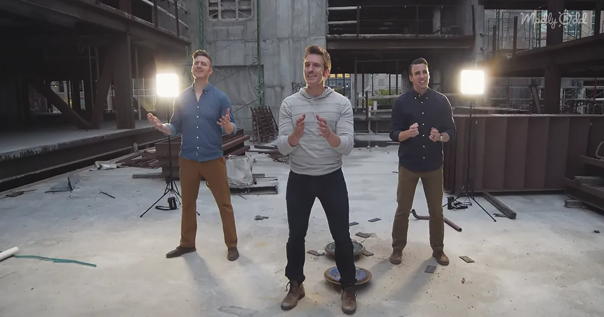 Three men in casual attire stand on a construction site, clapping and smiling under bright lights.
