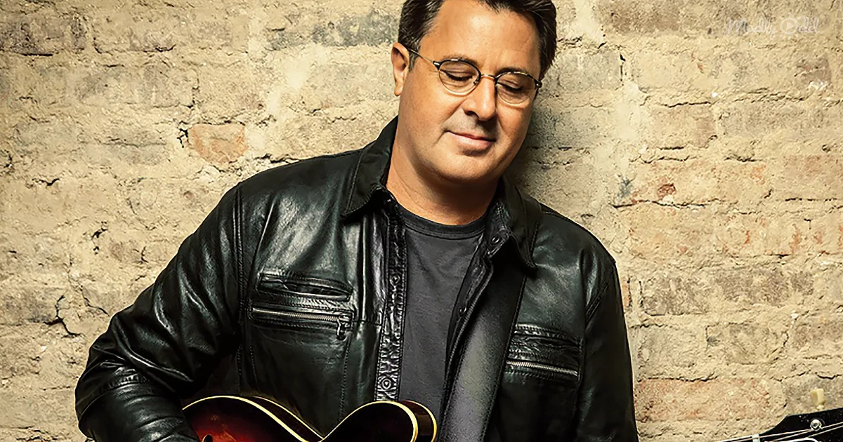 A man in a black leather jacket holds a guitar, leaning against a brick wall.