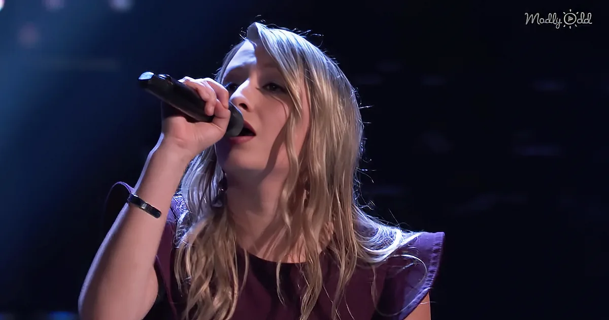 A person with long blonde hair sings into a microphone on stage, wearing a dark top.
