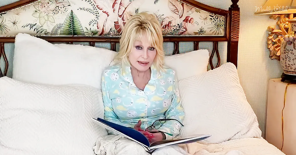 Dolly Parton sits on a bed in pajamas, holding a book and glasses, with a floral headboard and white pillows.