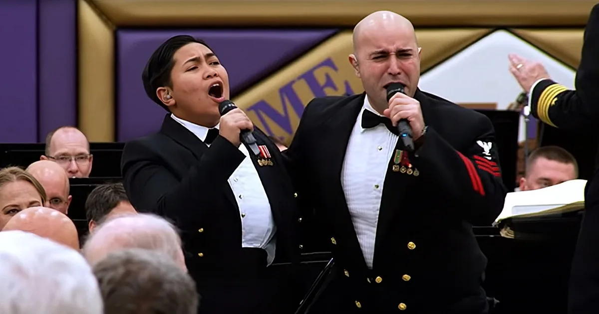 Two uniformed individuals sing into microphones in front of an audience, likely at a formal event.