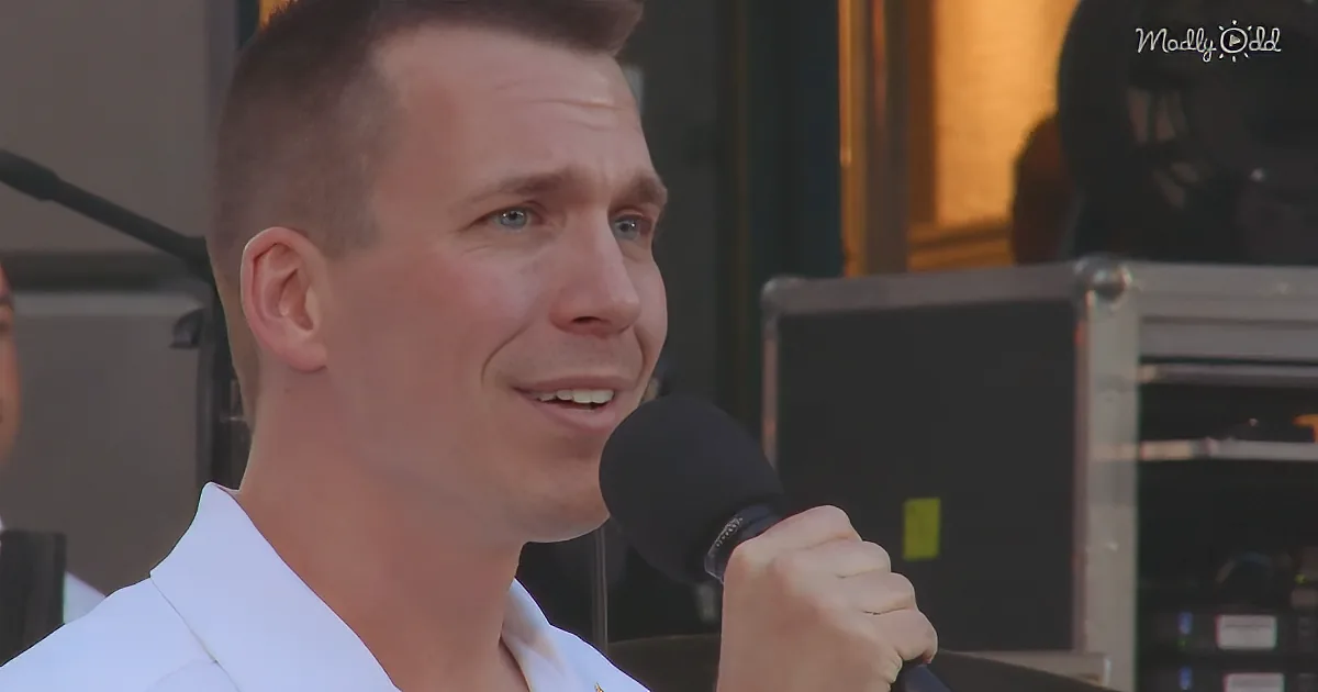 A man in a white shirt holds a microphone, speaking with a slight smile, in front of stage equipment.