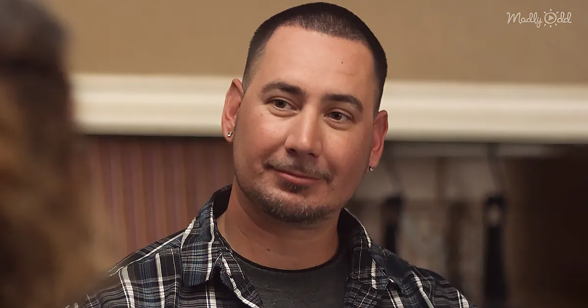 A man with short hair, earrings, and a plaid shirt is smiling and looking to the side.