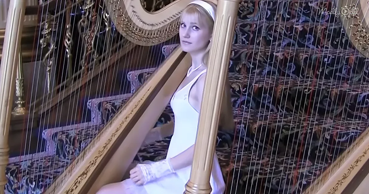A woman in a white dress sits beside a golden harp, looking directly at the camera.