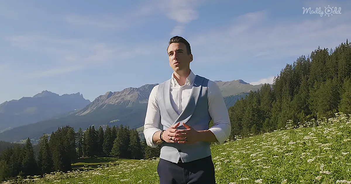 A man in a white shirt and light blue vest stands in a field of wildflowers, with a backdrop of mountains and trees.