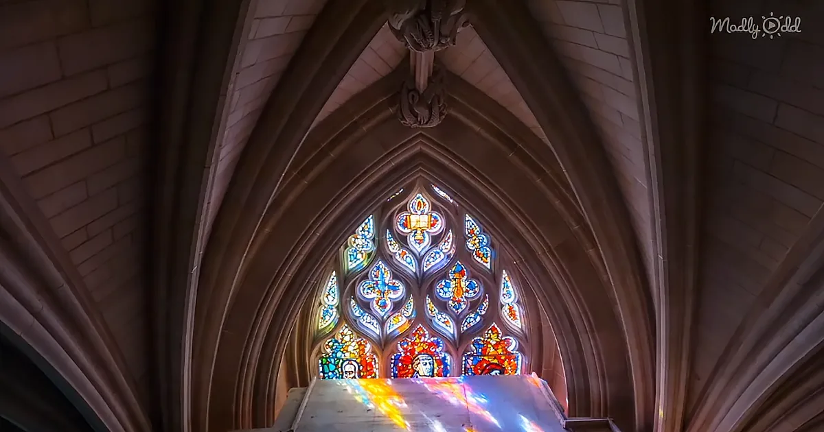 A grand stained glass window with vibrant colors and intricate designs is framed by ornate stone arches in a cathedral.