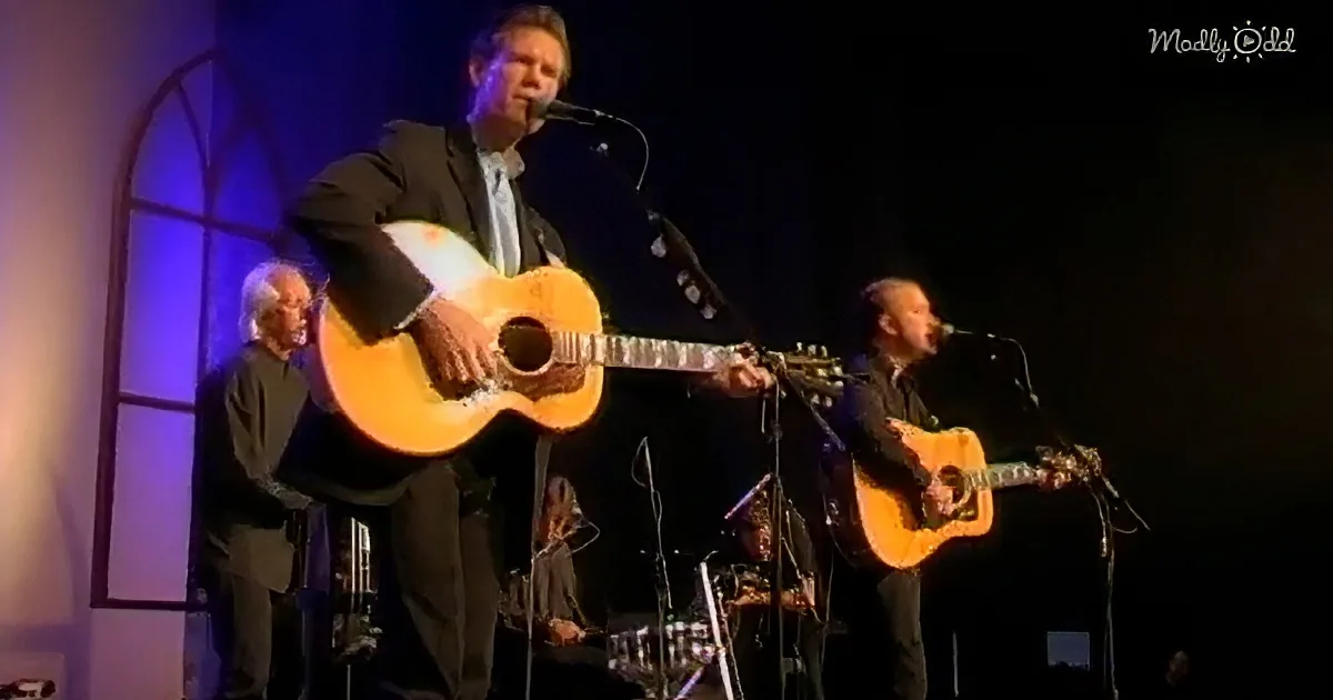 A man in a suit plays an acoustic guitar on stage, accompanied by other musicians.